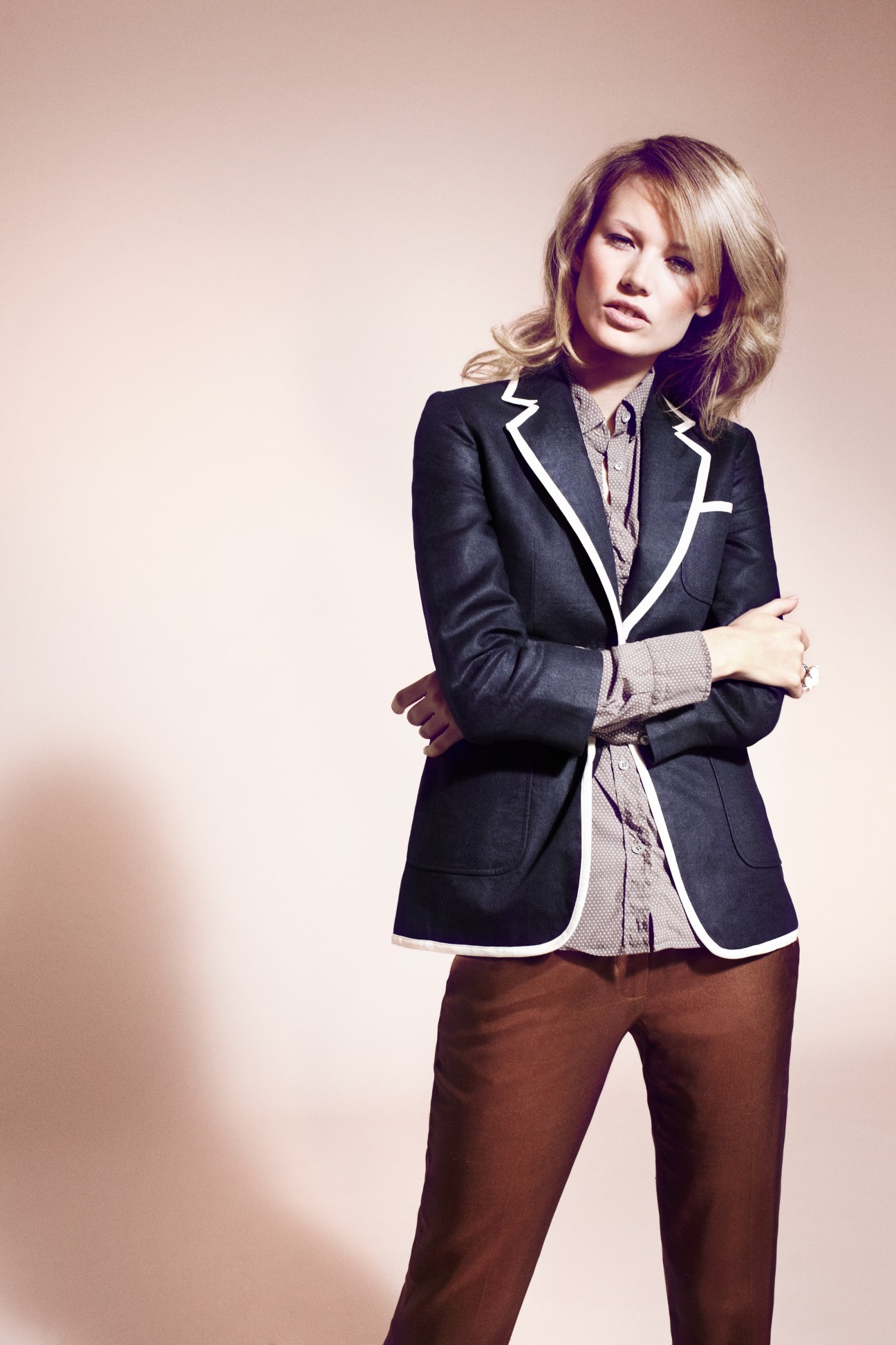 Woman wearing a navy blazer with white trim, patterned shirt, and brown trousers against a neutral background.