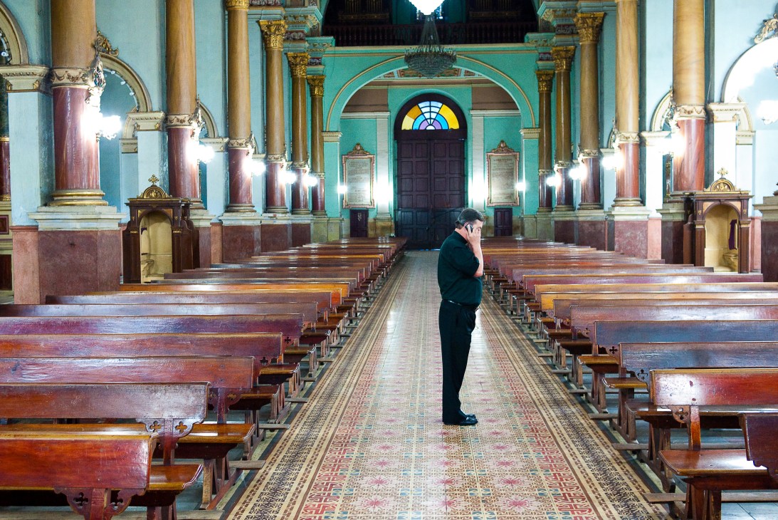 Padre Dario Echeverri stands alone in a ornate parish church with turquoise and gold columns.