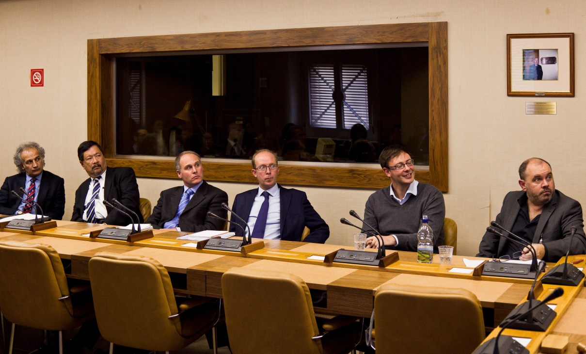 Six men seated at a long wooden table with microphones during a formal meeting in an institutional setting.