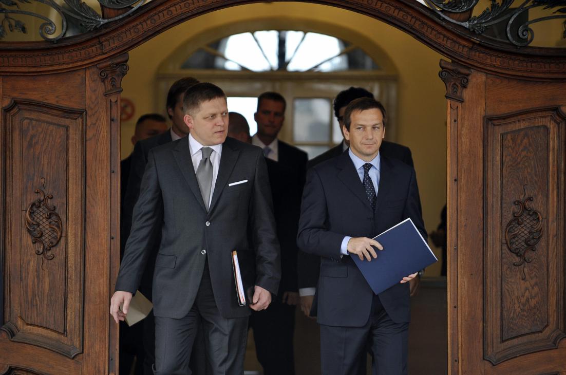 Two men in dark suits walk through ornate wooden doors, one holding a folder, with other officials visible behind them.