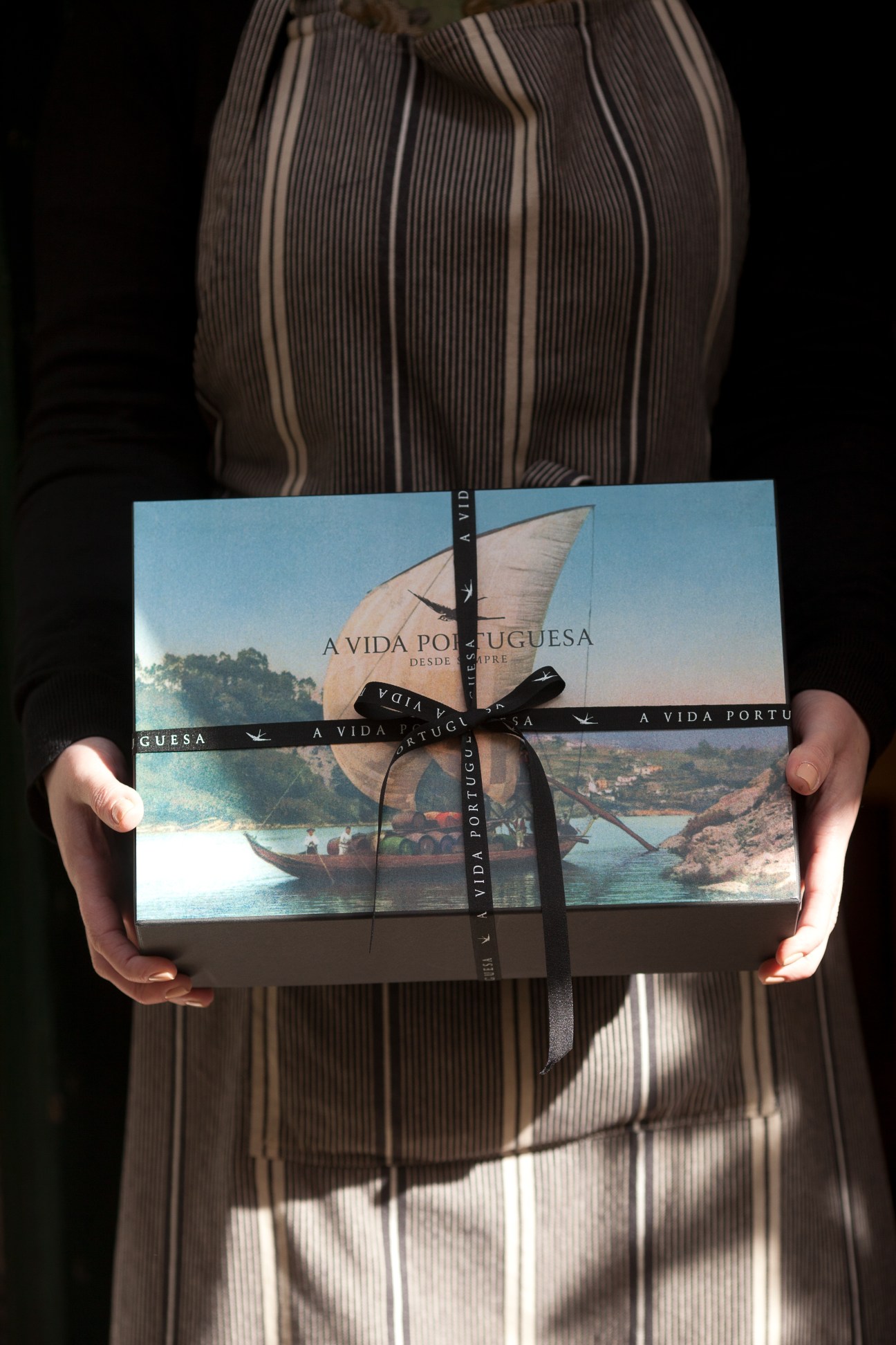 A Vida Portuguesa gift box tied with black ribbon, held by hands wearing striped clothing.