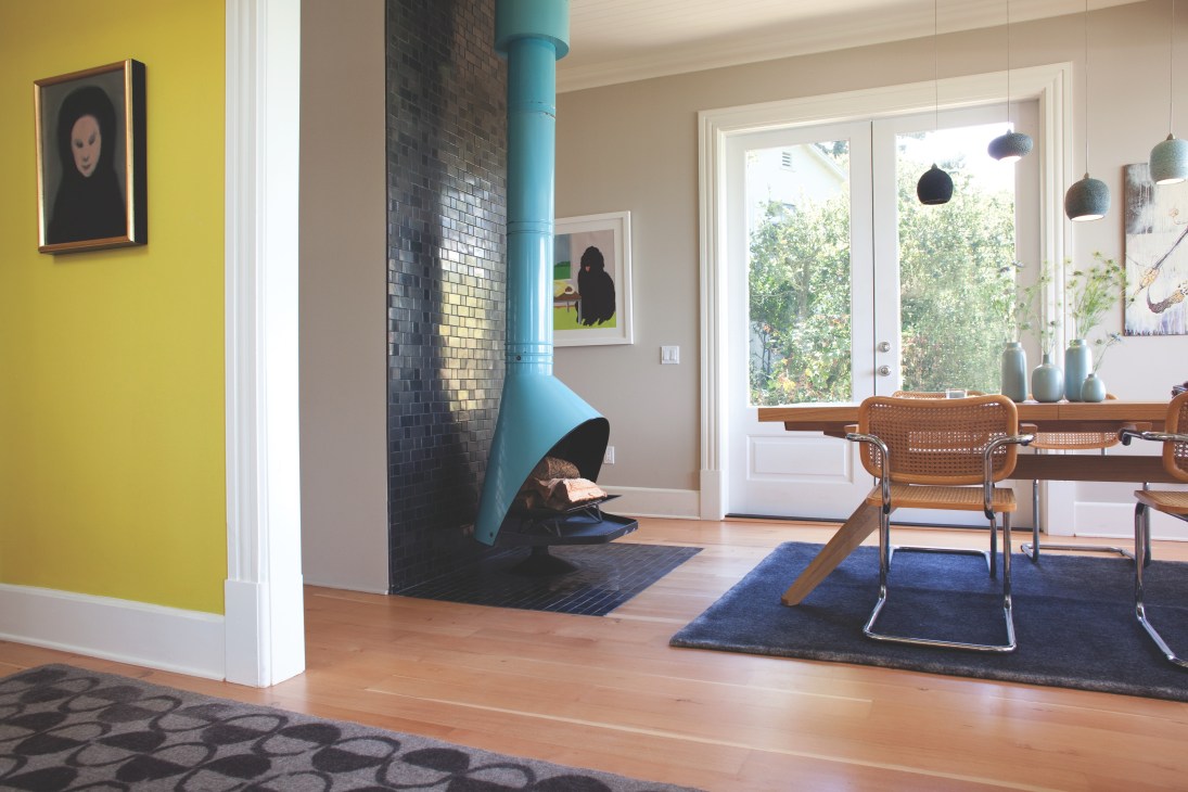 Dining room with turquoise hanging fireplace, yellow accent wall, wooden chairs, and black Heath ceramic tiles behind the fireplace.