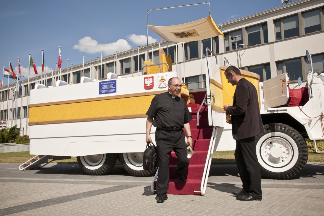A priest descends from a white and yellow replica Popemobile with red carpeted steps at an outdoor exhibition.