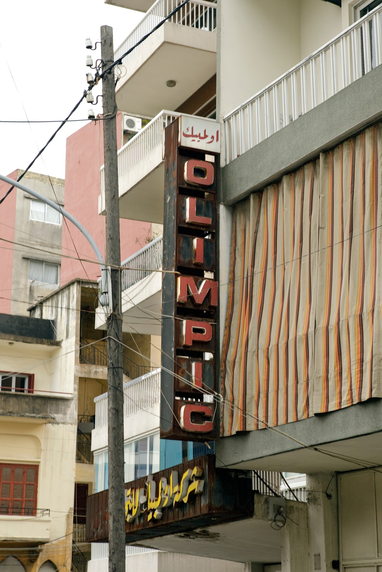 Vintage red neon sign on weathered pole in front of multi-story apartment building with Arabic text and 1970s design.