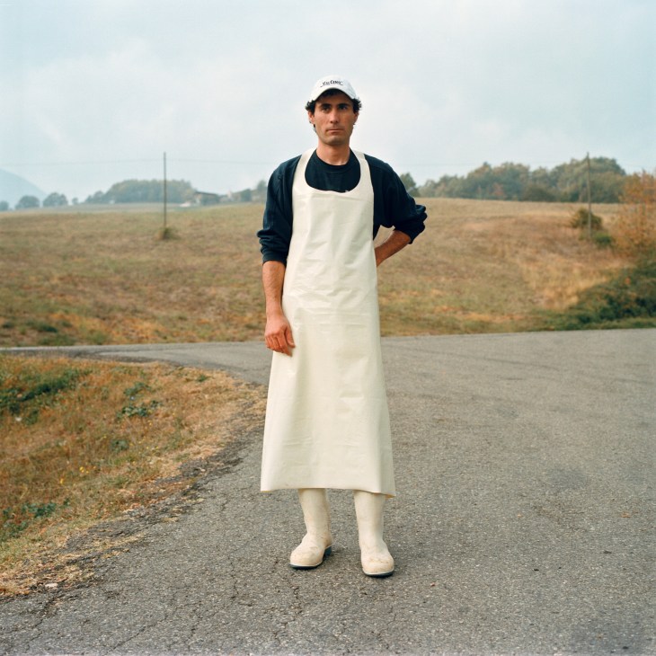 Cheesemaker Andrea Biolzi standing on a rural road wearing a white apron and cap, with fields behind him.