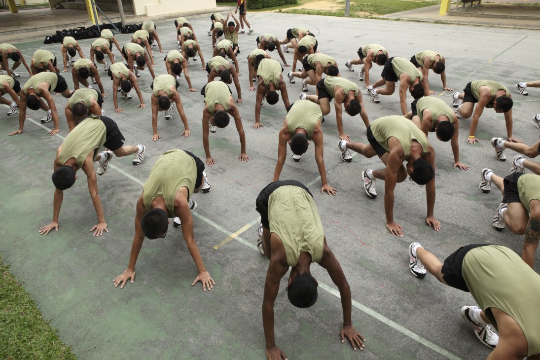 Multiple military recruits performing push-ups in uniform on an outdoor training ground at Palau Tekong.