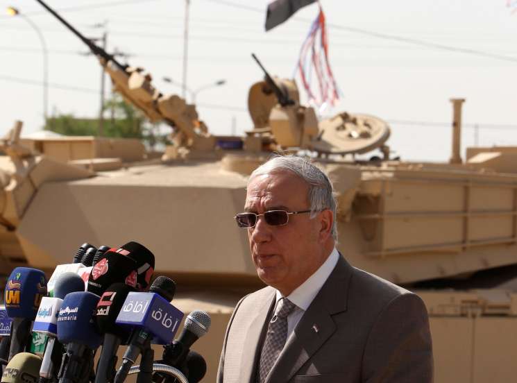 Man in suit and sunglasses speaks at microphone podium with Iraqi flag and military vehicle in background.