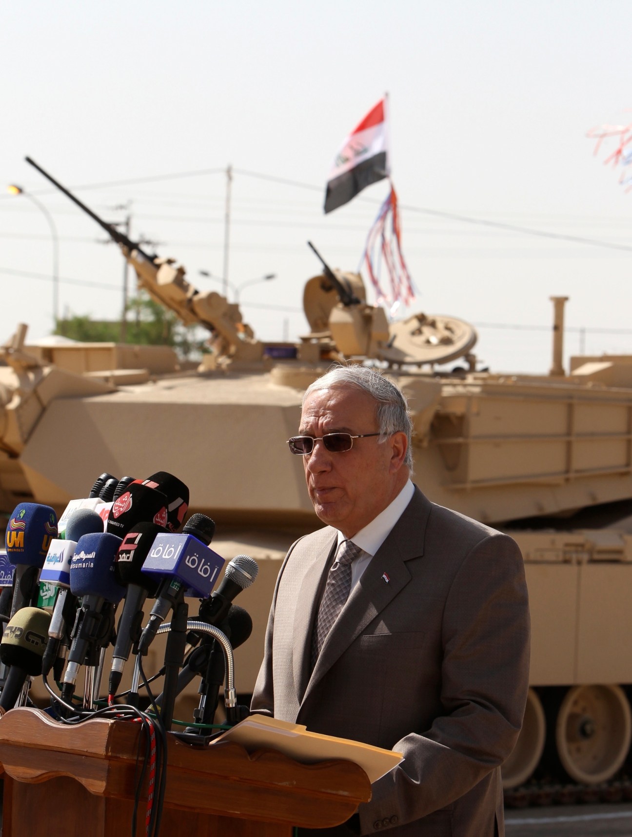Man in suit and sunglasses speaks at microphone podium with Iraqi flag and military vehicle in background.