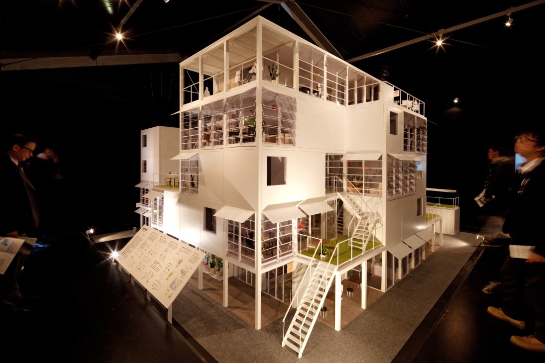 Illuminated architectural model of a multi-story community housing project with shared spaces, viewed by visitors in an exhibition setting.