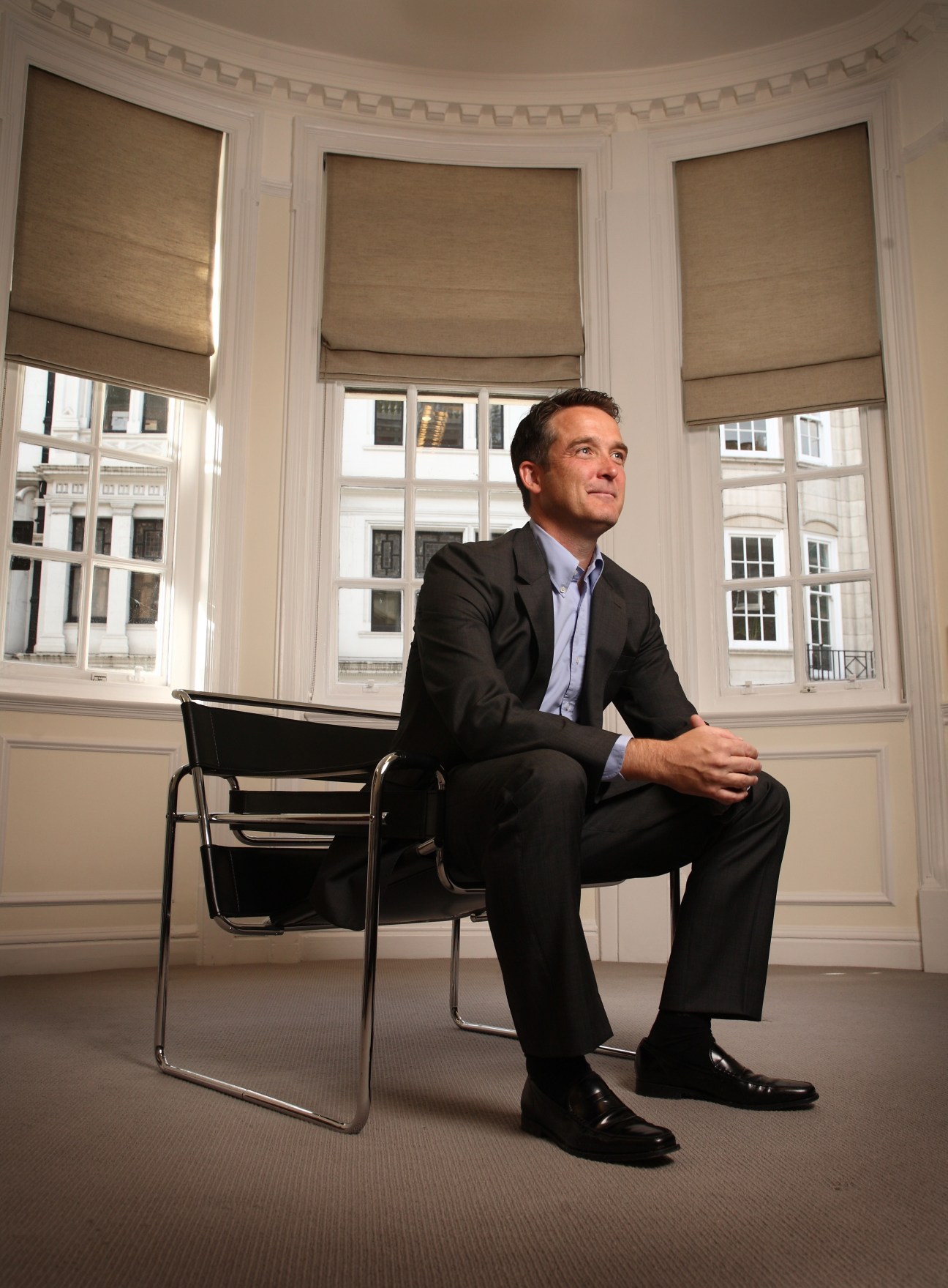 Man in dark blazer and light blue shirt sitting on modern chair in elegant room with large windows overlooking city streets.