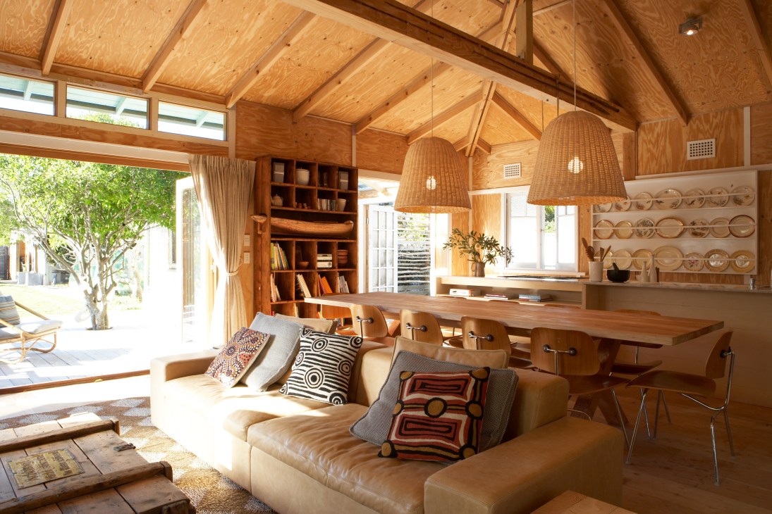 Wooden beach house interior with open-plan living and dining area, leather sofa, wicker pendant lights, and frangipani tree visible through glass doors.