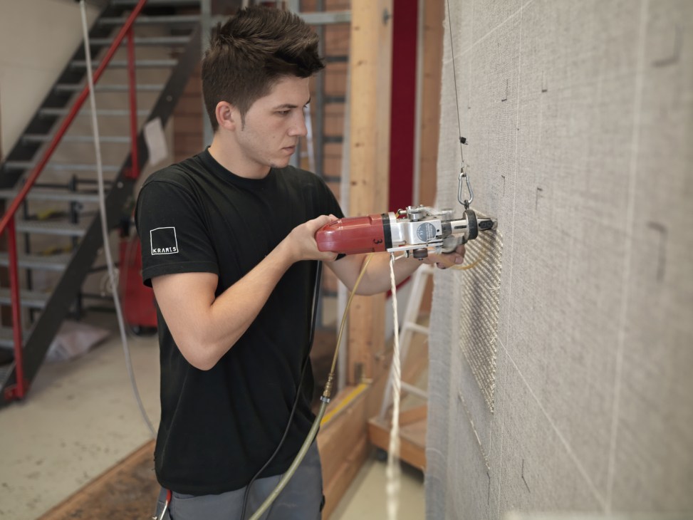 Young man using a hand-tufting drill instrument on a carpet mounted in the Kramis workshop.