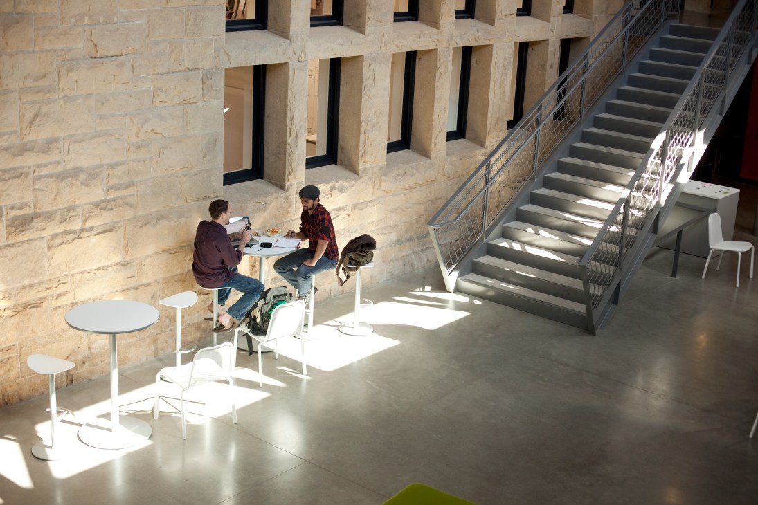 Two students work at a table in a bright, minimalist interior space with stone columns and stairs.