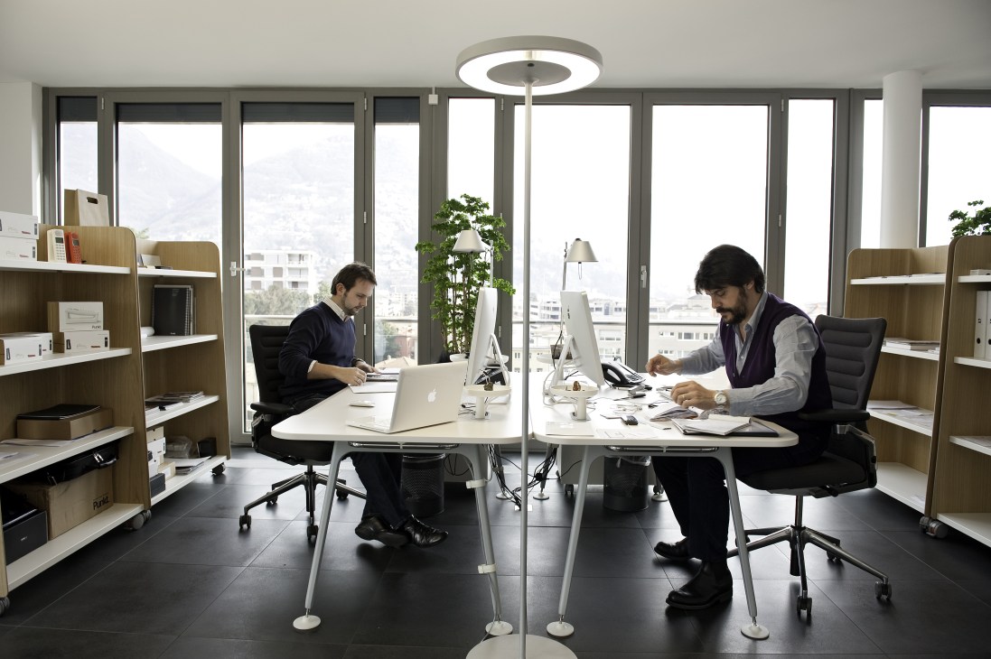 Two employees work at desks in a modern office with floor-to-ceiling windows overlooking Lake Lugano and mountains.
