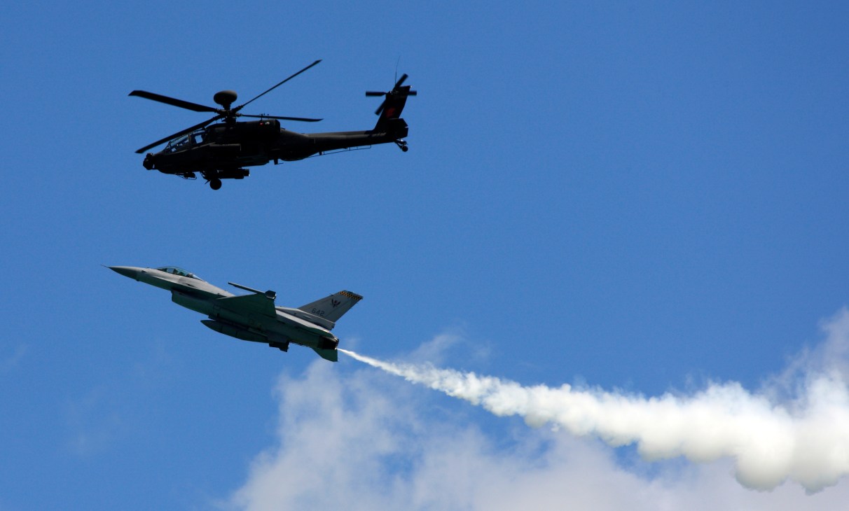 AH-64 Apache helicopter flying above an F-16 fighter jet with afterburner engaged against blue sky.