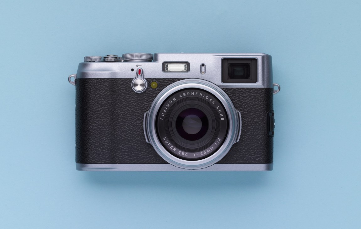 Fuji FinePix X100 compact digital camera with silver top plate and black leather body on blue background.