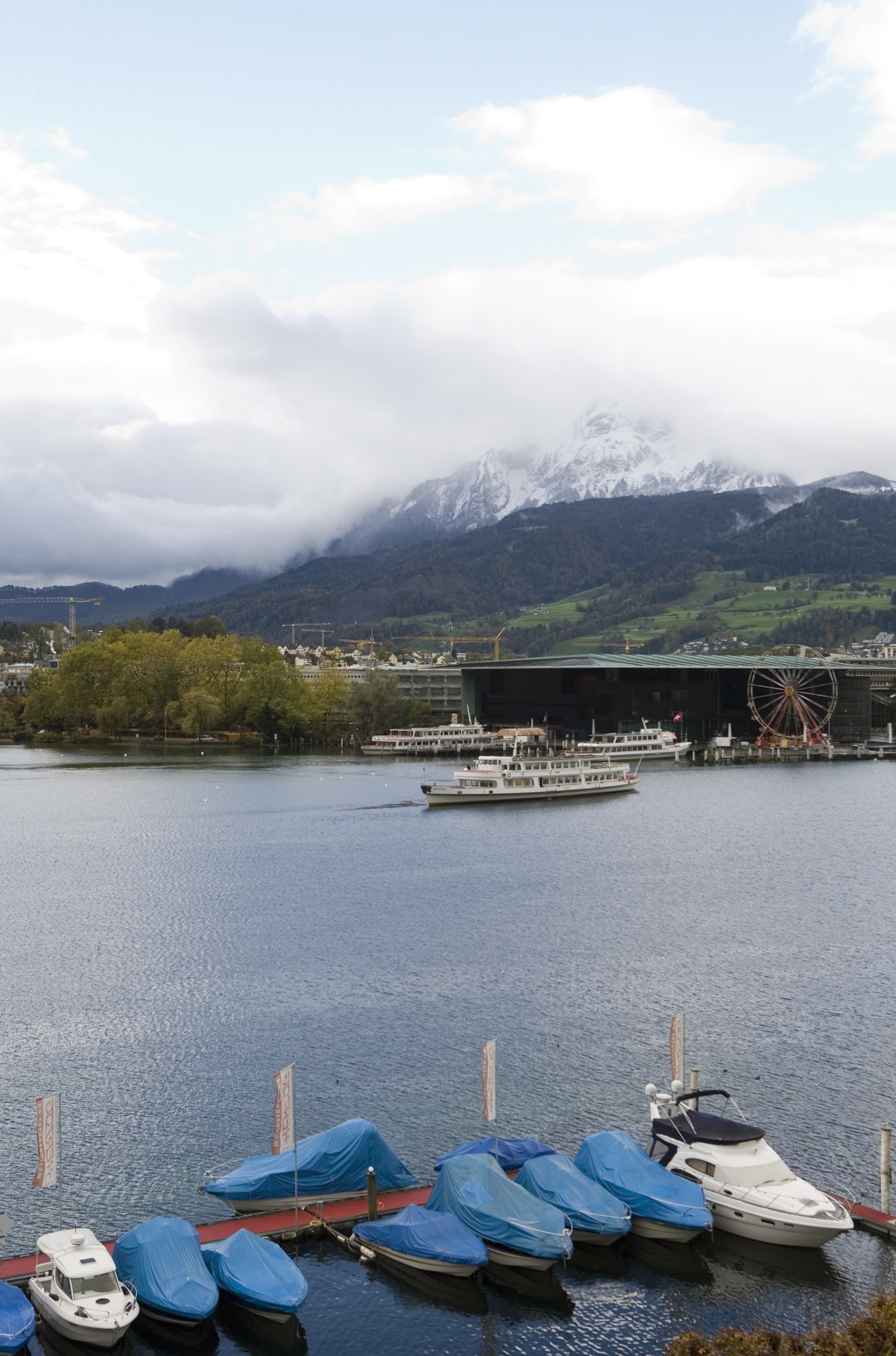 Lucerne lakeside with moored boats, snowy Alps and mountains in background under cloudy sky.