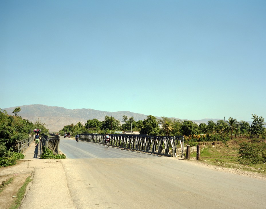 A metal bridge with white railings spans across a road on Haiti's outskirts, with mountains and vegetation visible in the background.