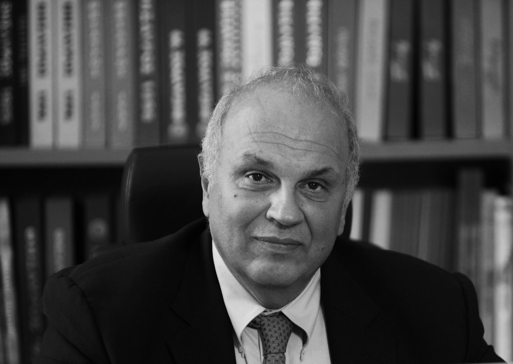 Black and white portrait of Spiro Pollalis, a man in a dark suit and tie seated in front of bookshelves.