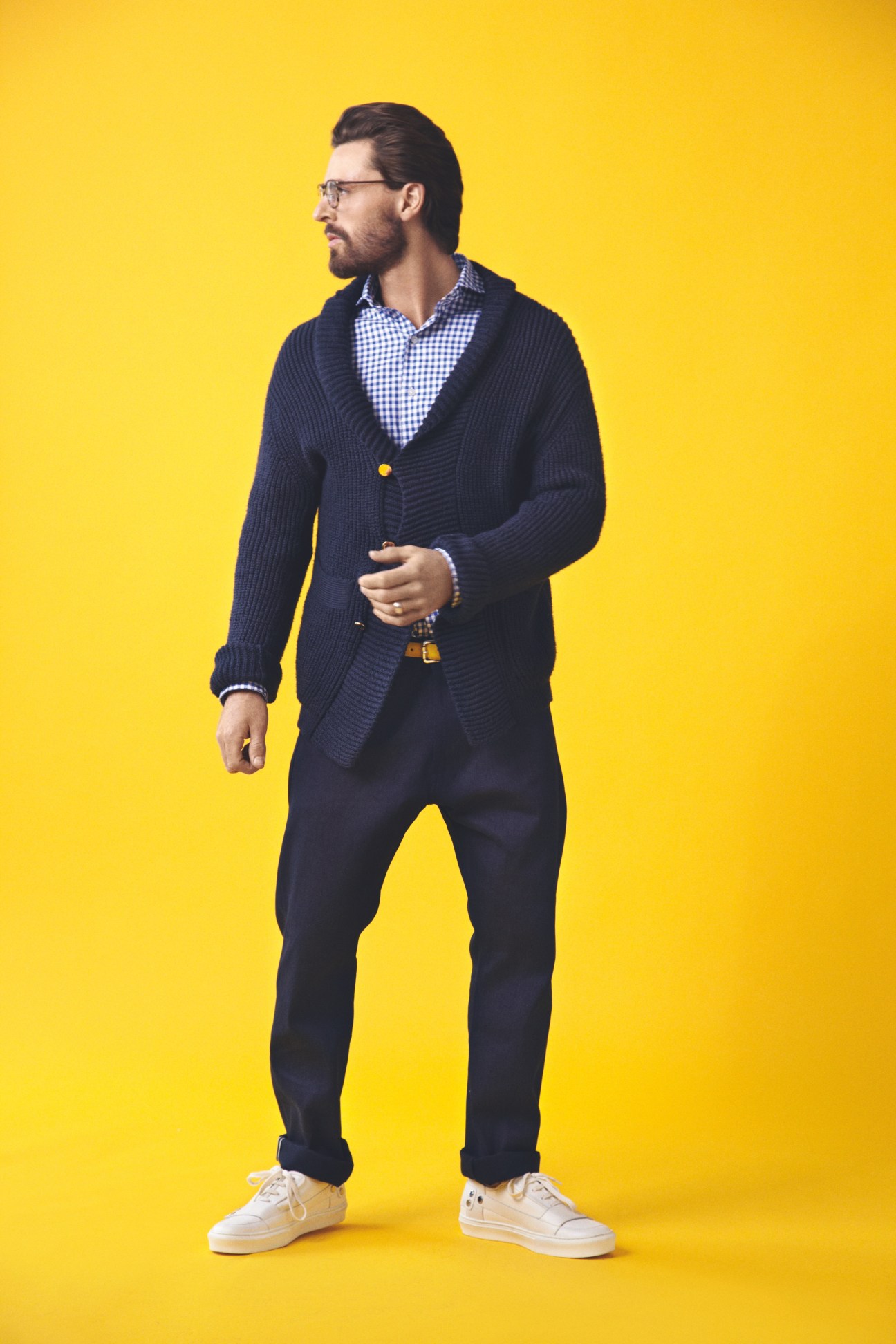 Man wearing navy cardigan, blue checked shirt, dark trousers, and white sneakers against yellow background.