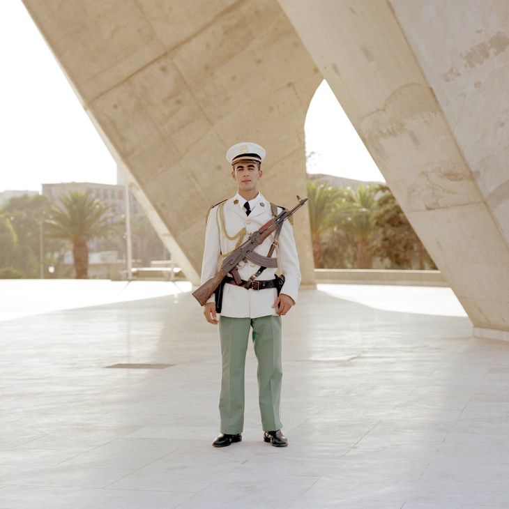 Bilayd Filak in white uniform and cap holding rifle at Martyrs' Memorial in Algiers.