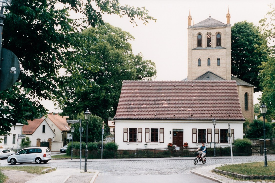 Wannsee village square with church, white building, parked car, and person on bicycle