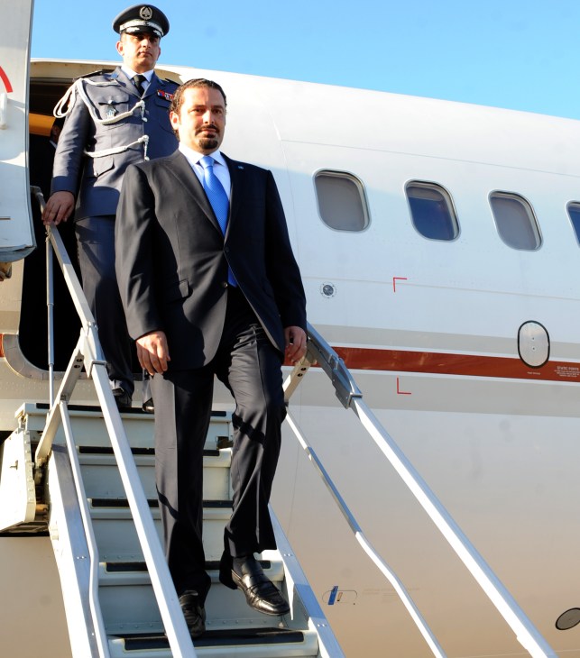 A man in a dark suit and blue tie descends airplane stairs accompanied by a uniformed military officer.