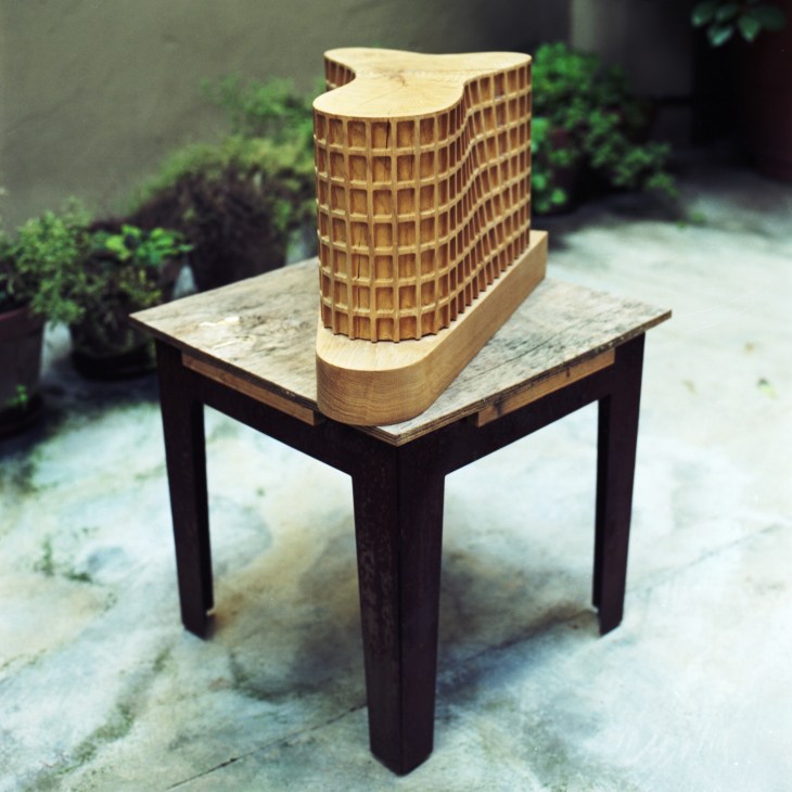 Wooden architectural model of a curved office building with gridded facade on a table in a garden setting.