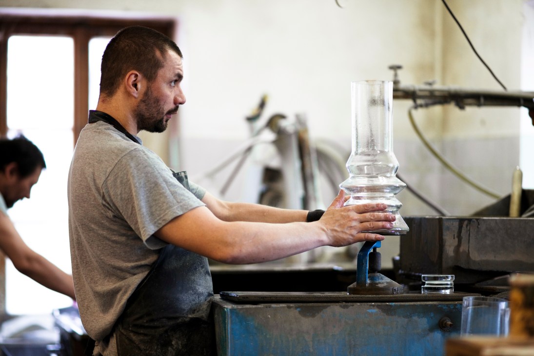 A glassmaker in the polishing workshop inspects a clear glass vessel for levelness.