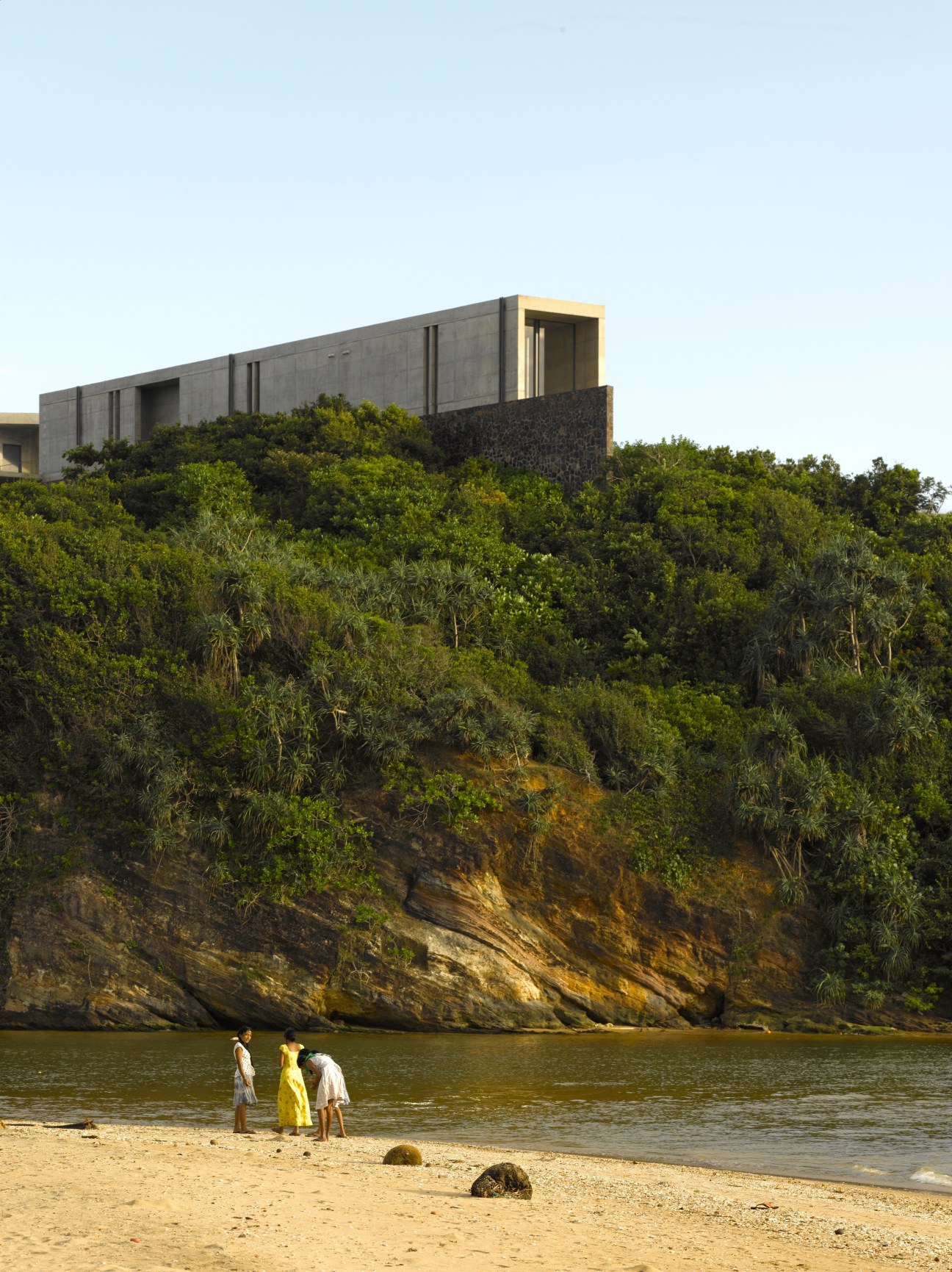 Tadao Ando's concrete house perched on a cliff above a beach with three people wading in the water below.