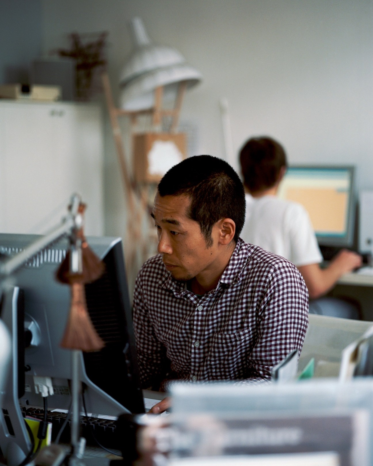Man in checkered shirt working at a desk in a design studio with colleagues and furniture models in the background.