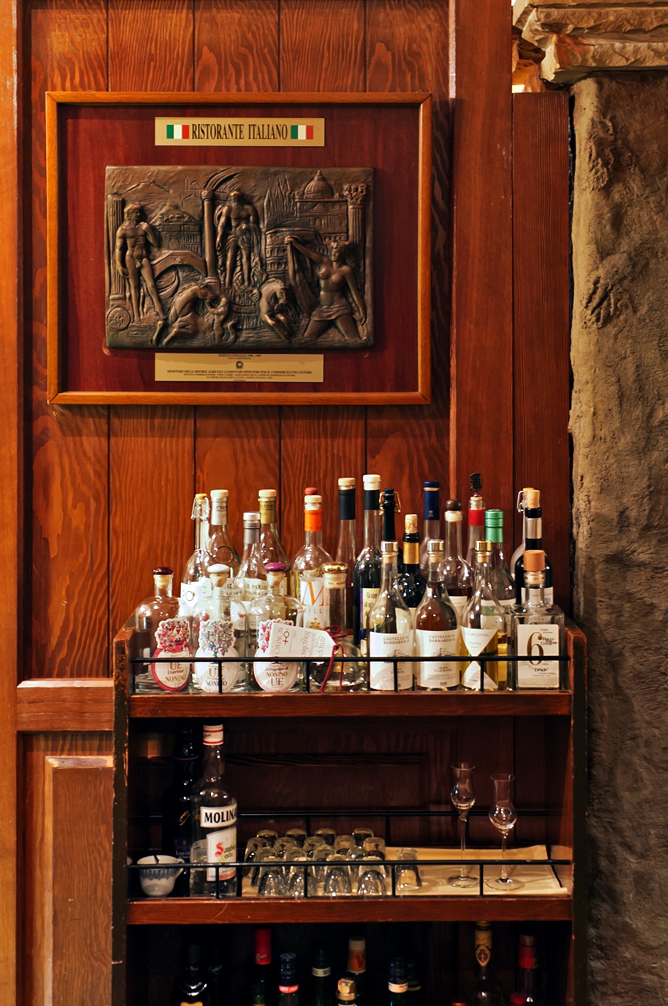 Wooden cabinet displaying various grappa bottles with decorative relief panel above at La Bisboccia restaurant.