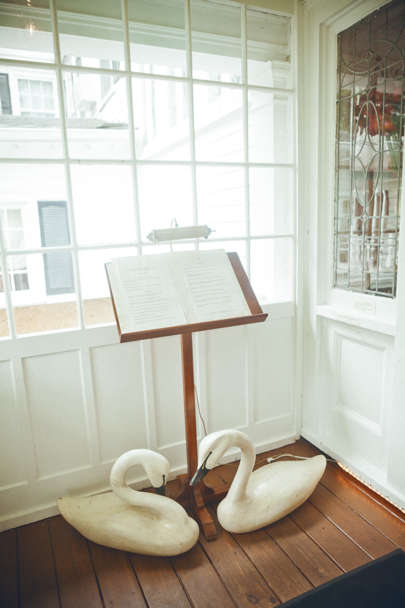 A wooden menu stand displays open pages beside two white carved swan decorations near a bright window.