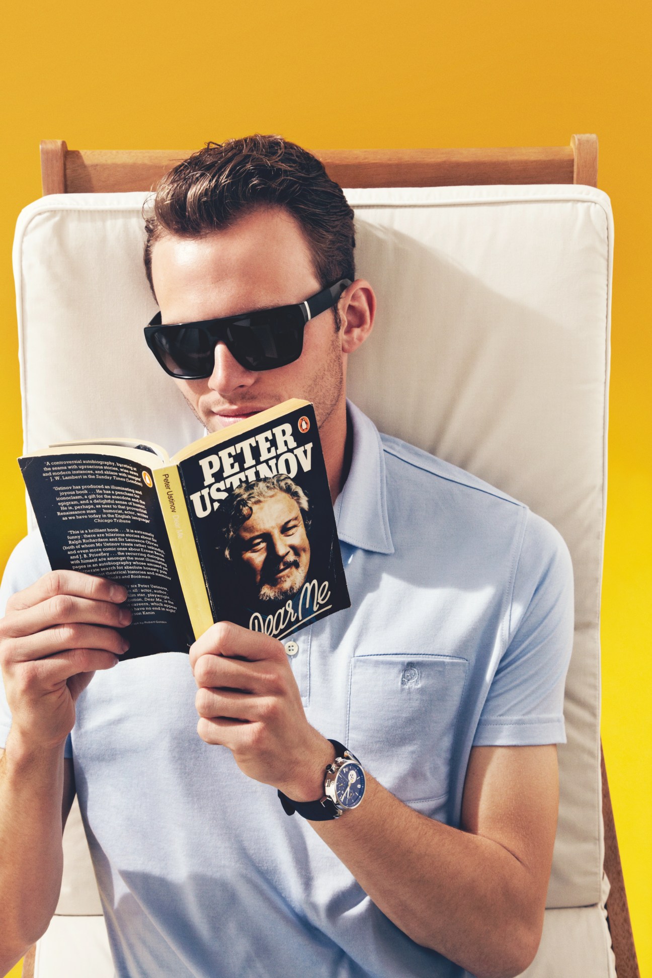 Man wearing Yves Saint Laurent sunglasses and Dunhill polo shirt reading a book against yellow background.
