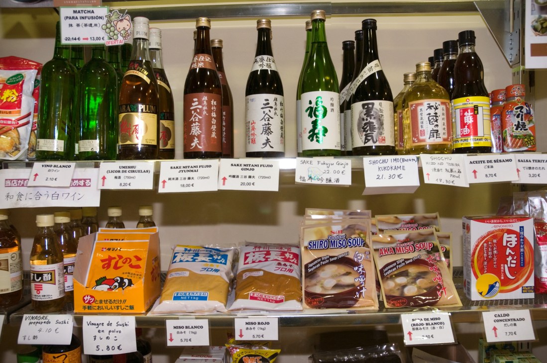 Shop display shelf with Japanese beverages including sake, shochu, and umeshu, plus packaged snacks and condiments with price labels.