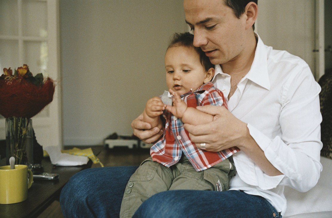 An Australian father in a white shirt holds his young son wearing a red plaid shirt indoors.