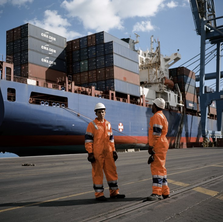 Two workers in orange safety suits and hard hats stand before a large container ship at DP World Sokhna port.
