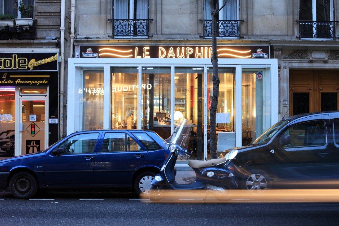 Le Dauphin restaurant storefront with neon signage on a Paris street lined with parked cars.
