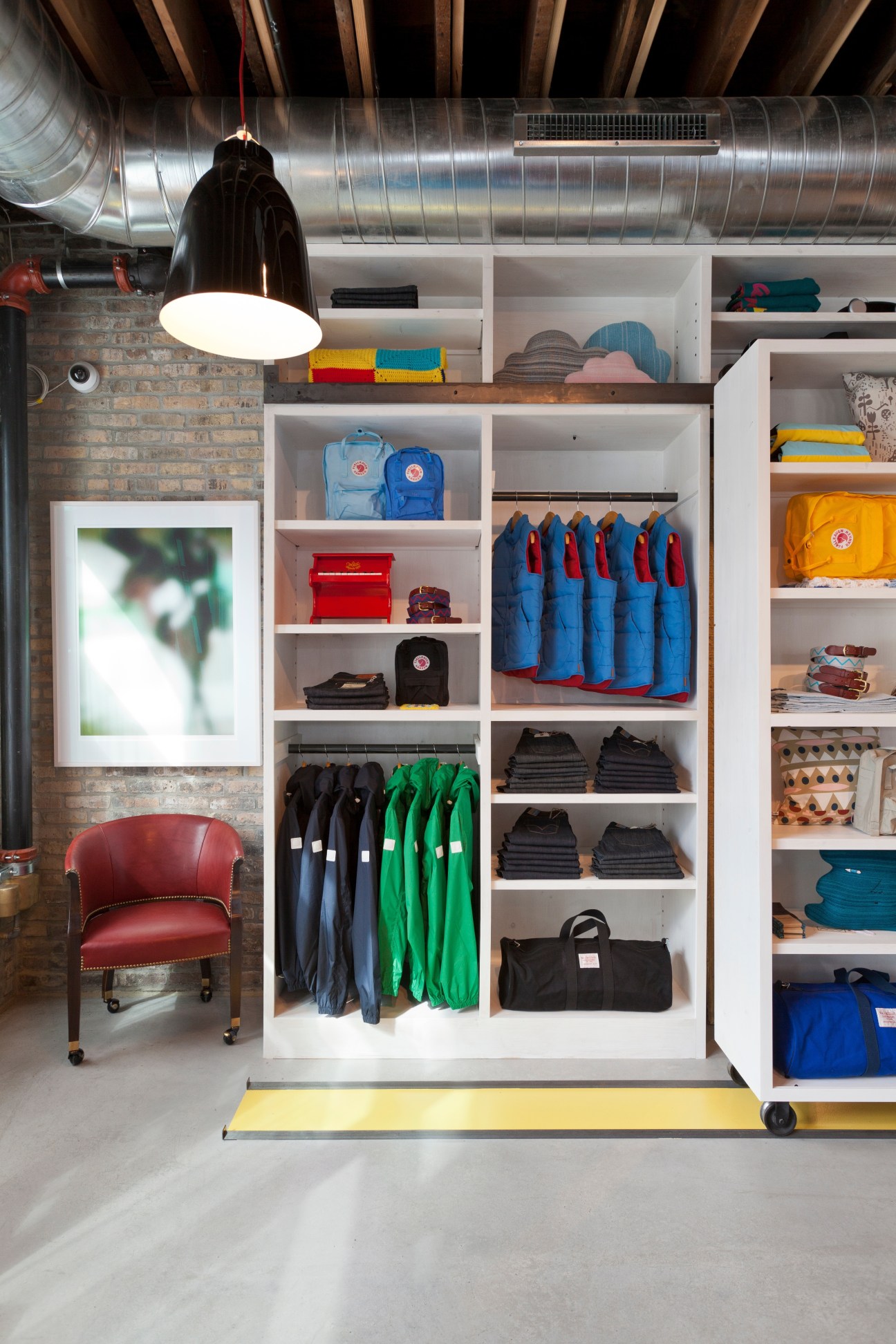 Askov Finlayson concept store interior with white shelving displaying colorful clothing, jackets, and accessories with exposed brick wall and pendant lighting.