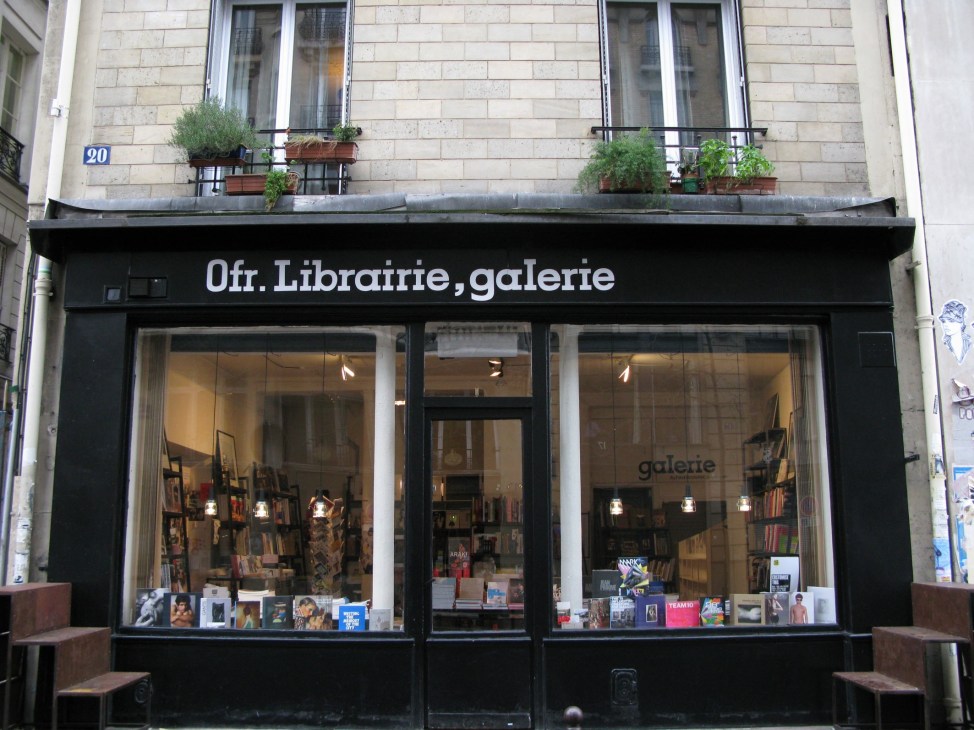 Storefront of Ofr. Librairie, galerie with black facade, large windows displaying books and magazines, and planters on upper level.