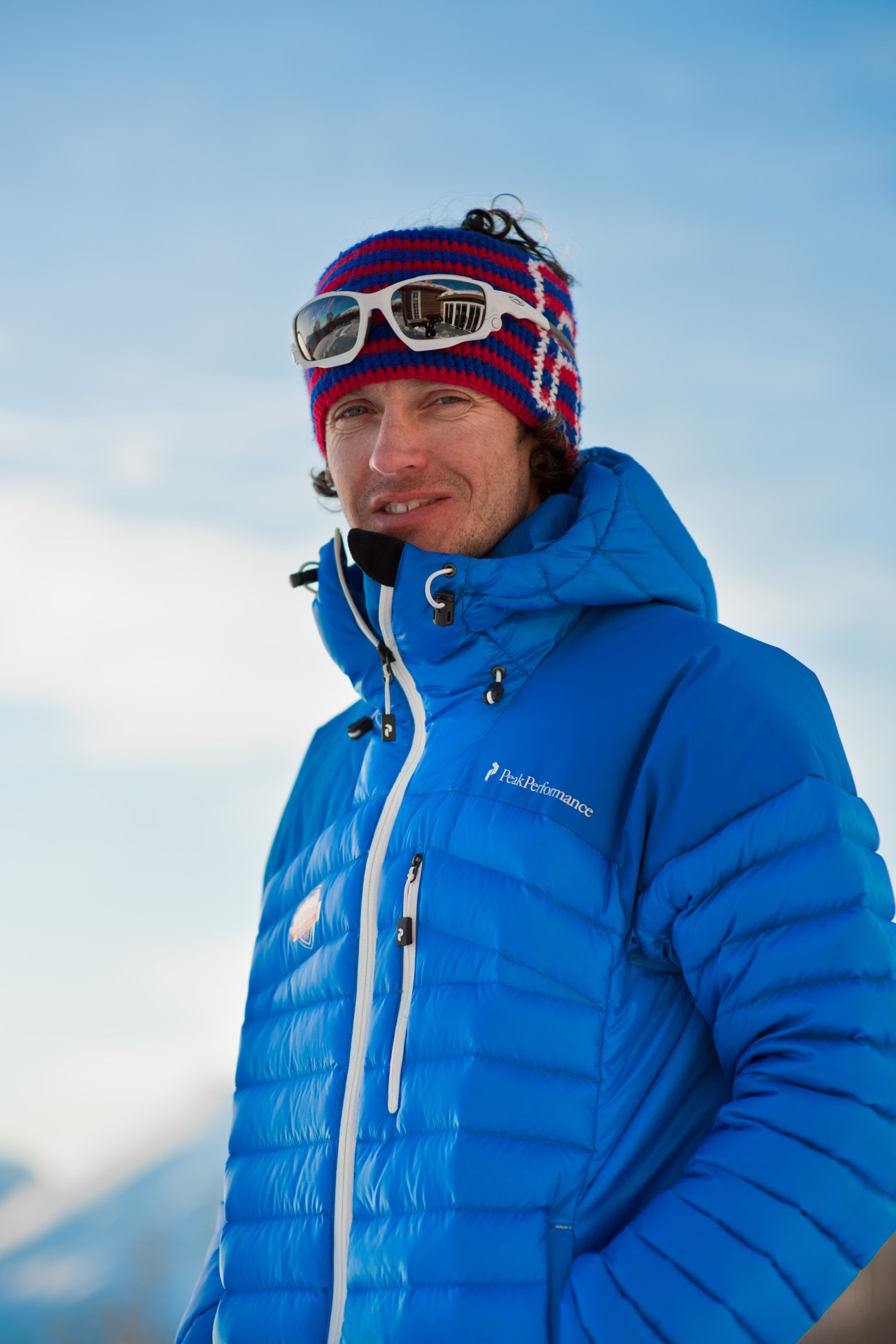 Man in blue puffer jacket and red-blue striped beanie with ski goggles smiles at camera against mountain backdrop.