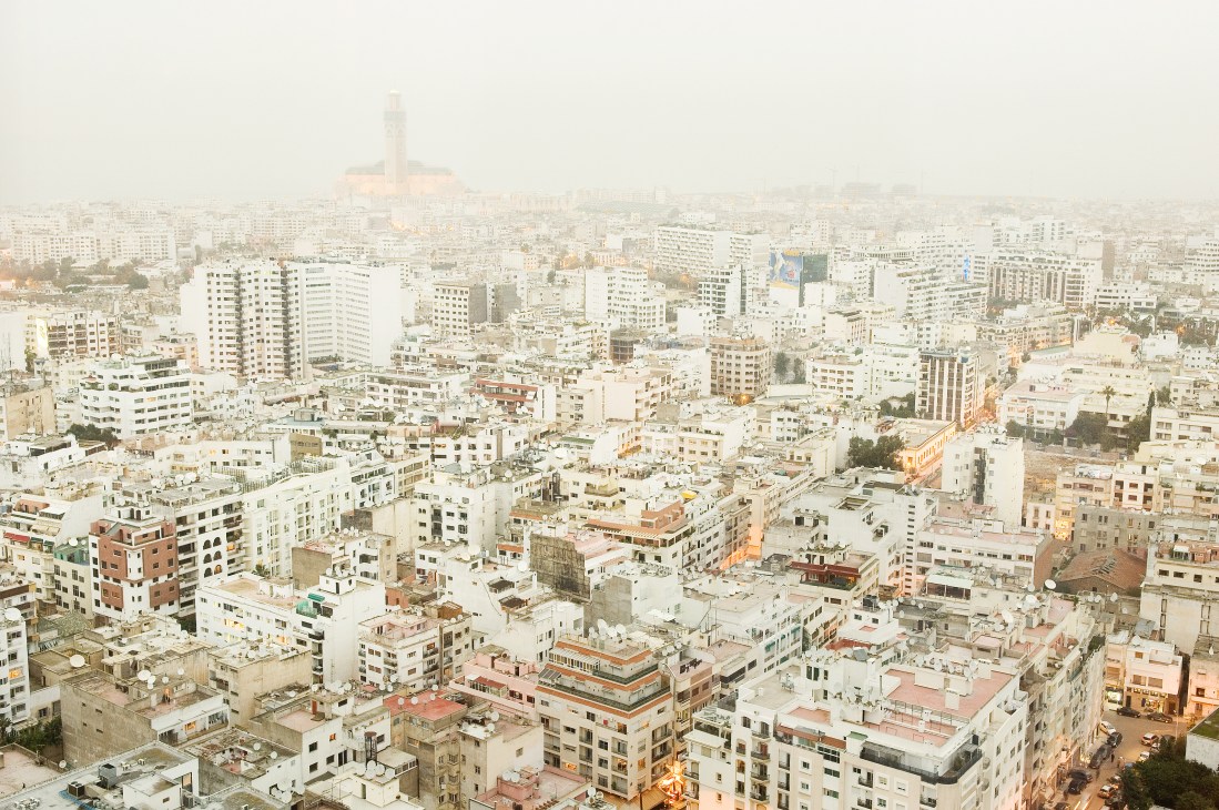 Aerial view of Casablanca showing dense residential buildings with Hassan II Mosque minaret in the distance under hazy sky.