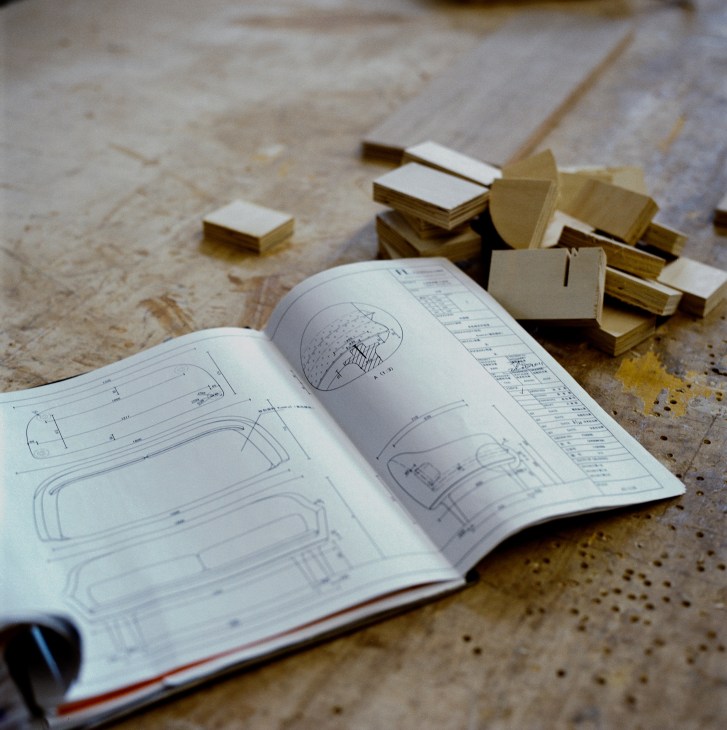 Open workbook with furniture design sketches and technical drawings on a concrete surface, surrounded by wood samples.