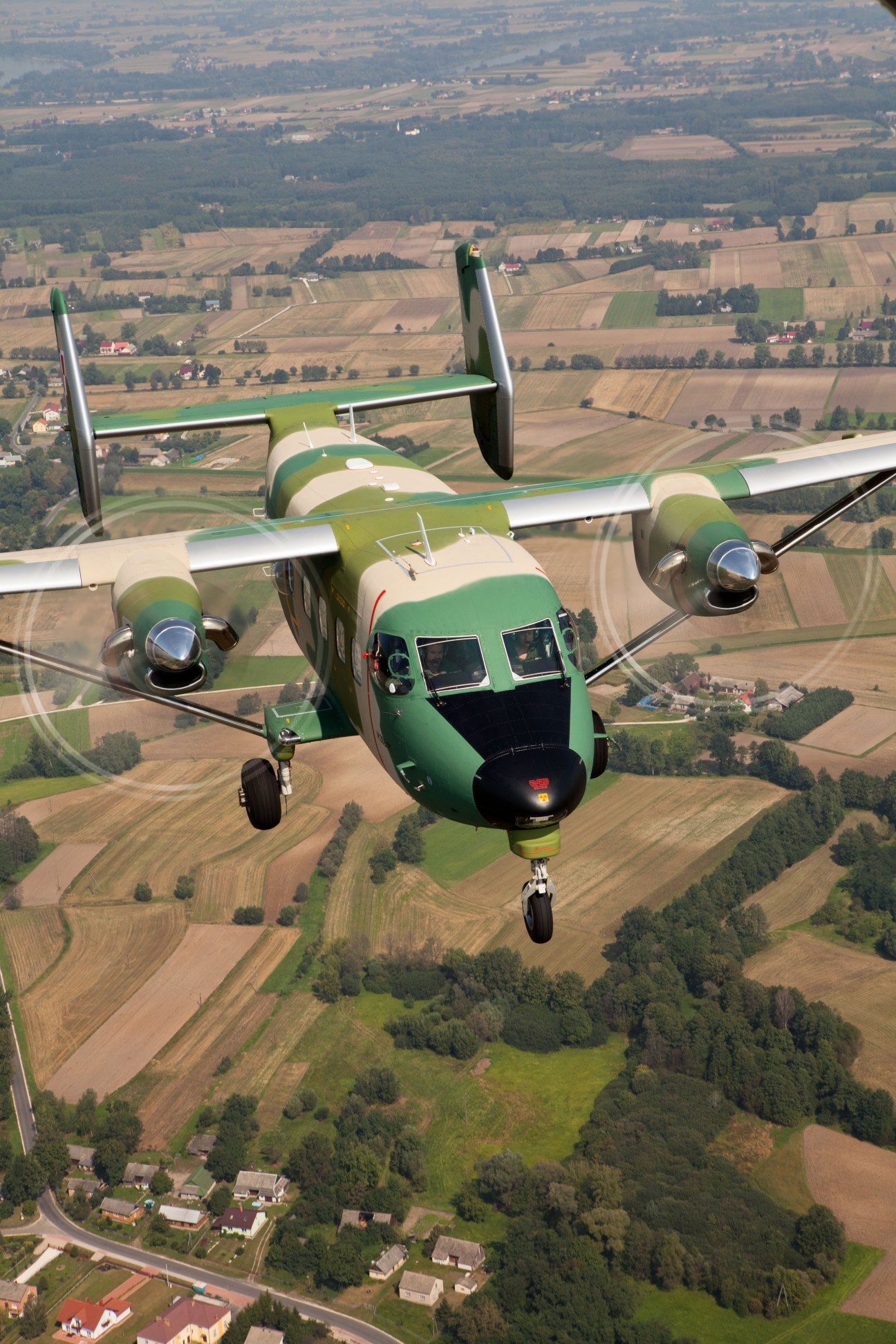 Green and white military transport aircraft in flight over patchwork farmland and forests.