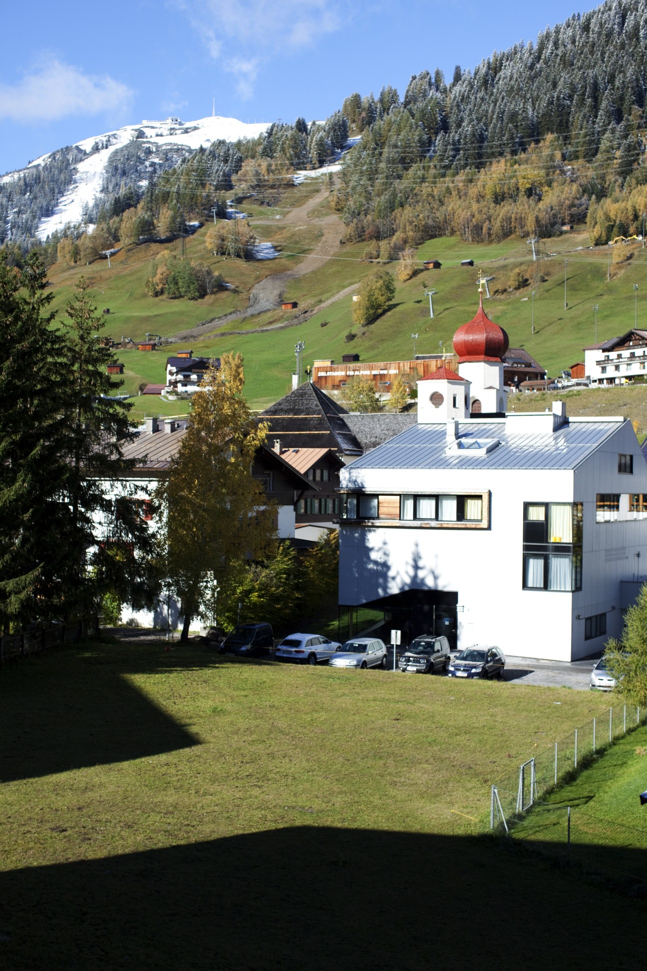 Modern white hotel building with red-domed chapel in Alpine valley surrounded by forested mountains and ski slopes.