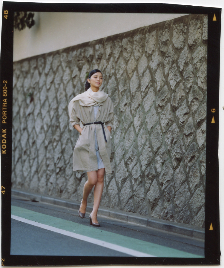 Model wearing a beige coat, Neil Barrett dress, and white stole walks past a textured stone wall in Tokyo.