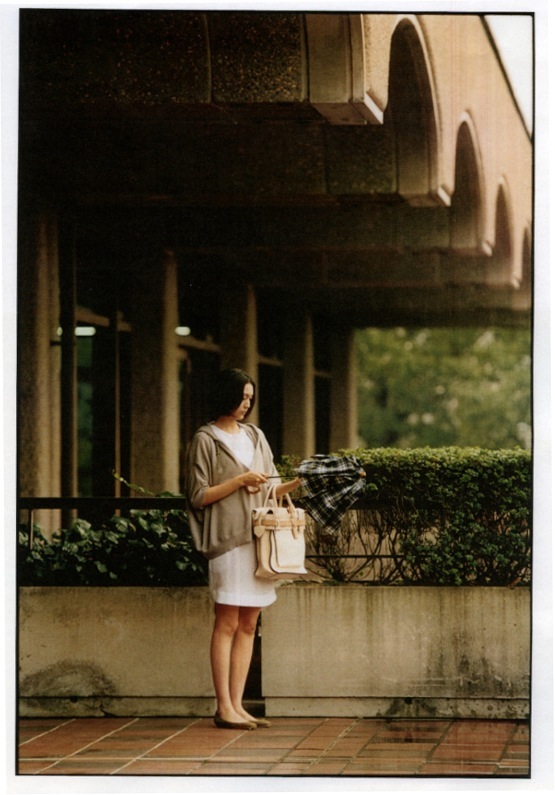 Woman in Reed Krakoff dress and Brunello Cucinelli hooded top standing under a covered colonnade holding an umbrella.
