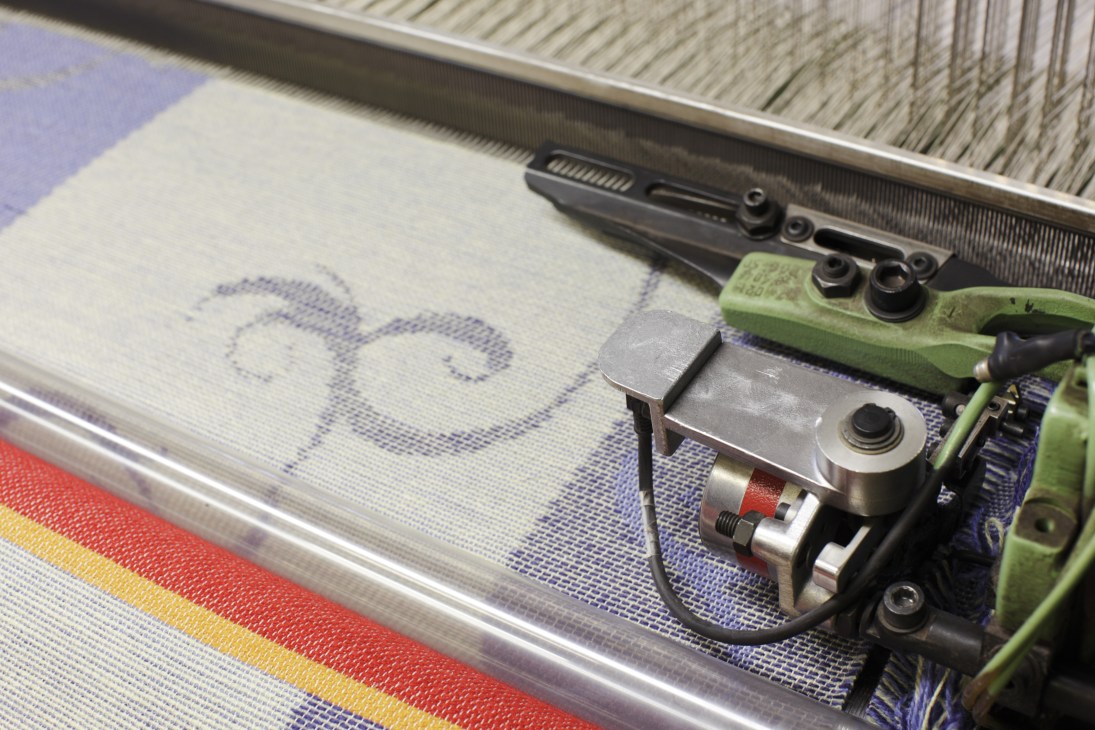 Close-up of a jacquard loom weaving Norwegian lambswool fabric with blue and colorful stripe pattern.