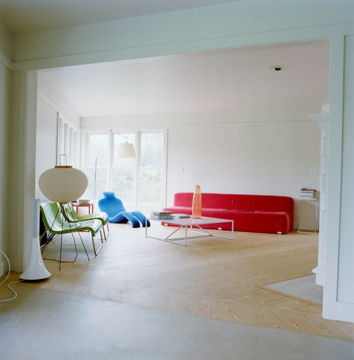 Minimalist living room with red sofa, green chair, paper lantern lamp and large windows overlooking nature.
