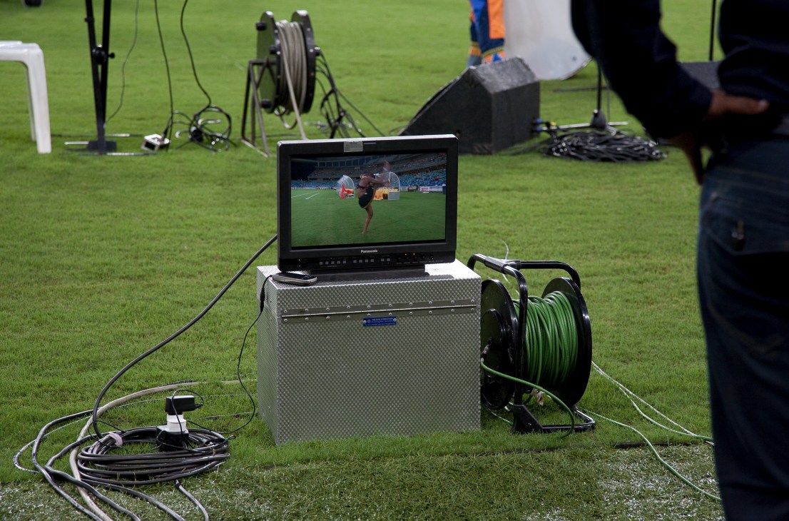 TV monitor displaying football match footage positioned on grass pitch surrounded by broadcasting cables and equipment.